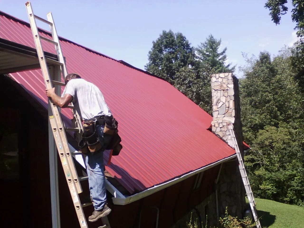 Roofer on a ladder beside a red metal roof and stone chimney — Soffit & Fascia Repair by Roofing Brothers in Sebree